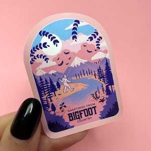K&ouml;nnte beinhalten: Ein rosa und blauer Aufkleber mit einem Bigfoot-Cartoon, der in einem Wald mit Bergen und einem Fluss im Hintergrund spaziert. Der Aufkleber lautet "Greetings from Bigfoot Country".