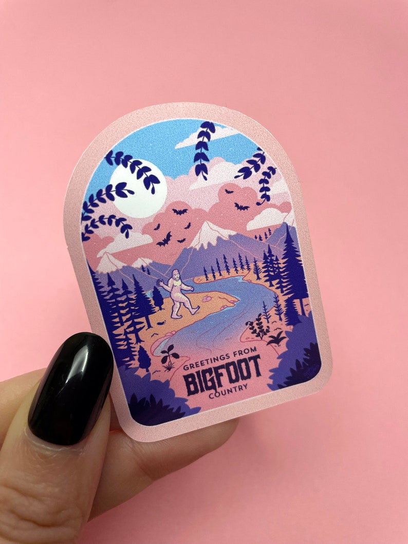 K&ouml;nnte beinhalten: Ein rosa und blauer Aufkleber mit einem Bigfoot-Cartoon, der in einem Wald mit Bergen und einem Fluss im Hintergrund spaziert. Der Aufkleber lautet "Greetings from Bigfoot Country".