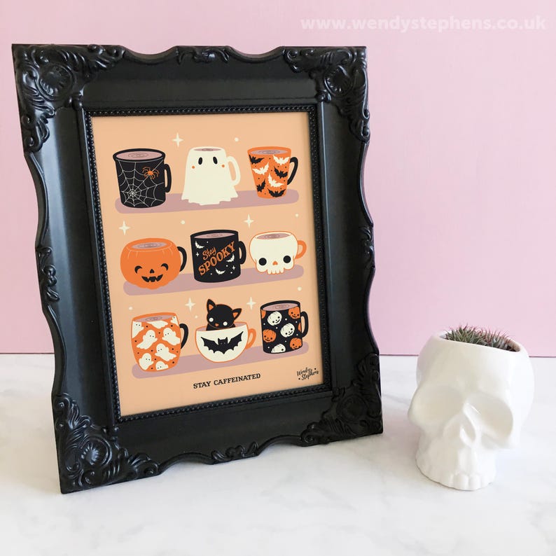 Op de afbeelding: Een ingelijste print met negen Halloween-thema mokken met verschillende ontwerpen, waaronder een spinnenweb, een spook en een pompoen. De print zit in een zwart, sierlijk frame, met een witte schedelvormige plantenbak ernaast. De tekst "Stay Caffeinated" staat onderaan.