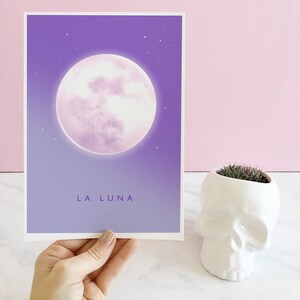 May include: A purple and white print with a large, full moon and the text "LA LUNA" at the bottom. The print is framed and hanging on a pink wall.