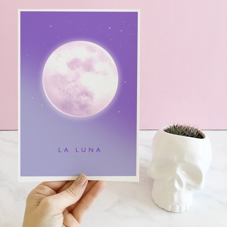 May include: A purple and white print with a large, full moon and the text "LA LUNA" at the bottom. The print is framed and hanging on a pink wall.