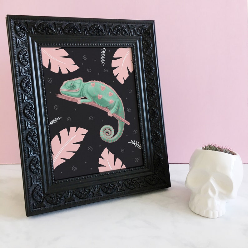 May include: A framed print of a green chameleon with pink hearts on its body, perched on a branch against a black background with pink tropical leaves.
