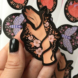 May include: A black sticker featuring a hand intertwined with a black snake, surrounded by pink flowers and foliage. The sticker has a dark background with additional floral and insect designs.