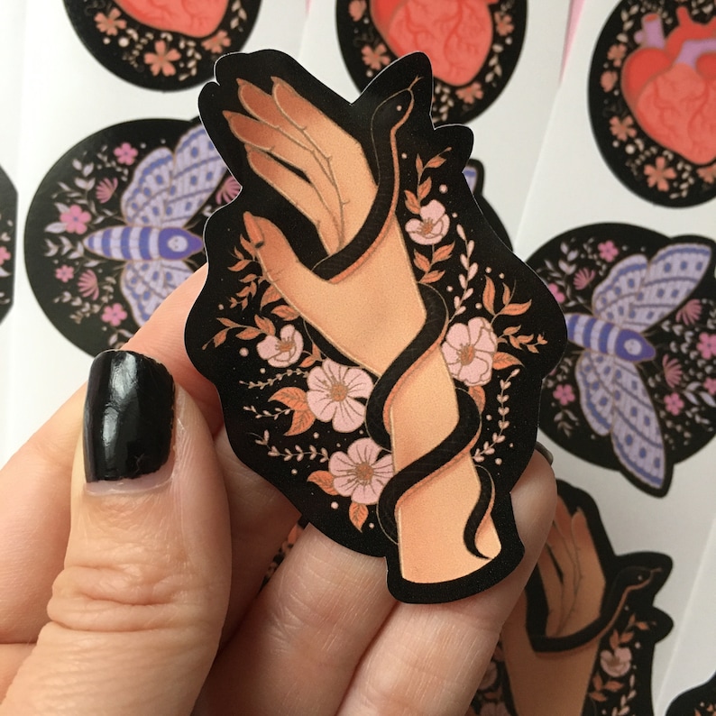 May include: A black sticker featuring a hand intertwined with a black snake, surrounded by pink flowers and foliage. The sticker has a dark background with additional floral and insect designs.