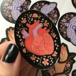 May include: Oval sticker featuring a stylized human heart in shades of red and pink, surrounded by floral elements in peach and white, set against a black background. Other stickers with moth designs are visible.