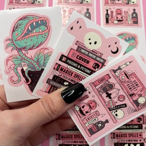 Puede incluir: Un conjunto de cuatro pegatinas rosas y negras con temas de Halloween espeluznantes. Las pegatinas incluyen una planta carnívora, un fantasma, una pila de libros con las palabras "Magick Spells" y una estantería con pociones, calaveras y calabazas.