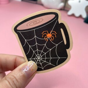 May include: A sticker featuring a black mug with a spider web design and an orange spider. The mug has a light brown rim and a handle. The sticker is on a pink background. Halloween themed.