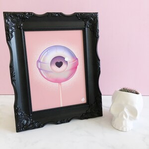 May include: A framed print of a pink and purple eyeball lollipop with a small heart in the center of the pupil. The lollipop is on a stick and is set against a pink background.