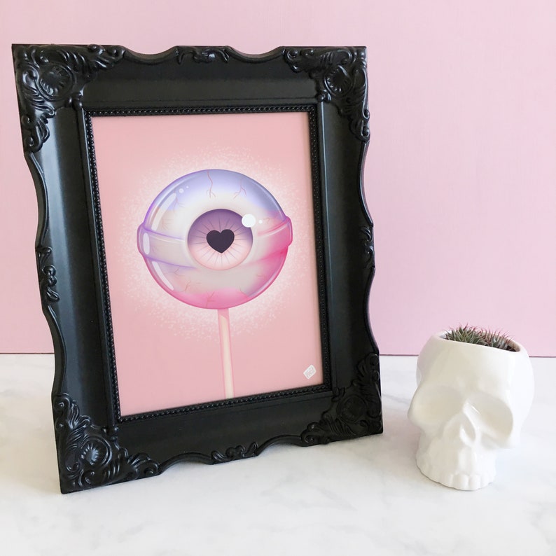 May include: A framed print of a pink and purple eyeball lollipop with a small heart in the center of the pupil. The lollipop is on a stick and is set against a pink background.