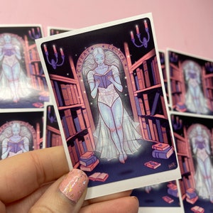 Puede incluir: Una pegatina con una figura fantasmal vestida con un vestido blanco leyendo un libro en un entorno de biblioteca. El texto "Caveat Lector" est&aacute; escrito en un c&iacute;rculo sobre la figura. La pegatina tiene un esquema de color morado y rosa.