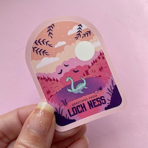 K&ouml;nnte beinhalten: Ein rosa und lilafarbener Aufkleber mit einer Cartoon-Illustration eines gr&uuml;nen Seeungeheuers im Loch Ness in einem See. Der Aufkleber hat einen rosa Bogen mit dem Text "Greetings from Loch Ness" in Wei&szlig;.
