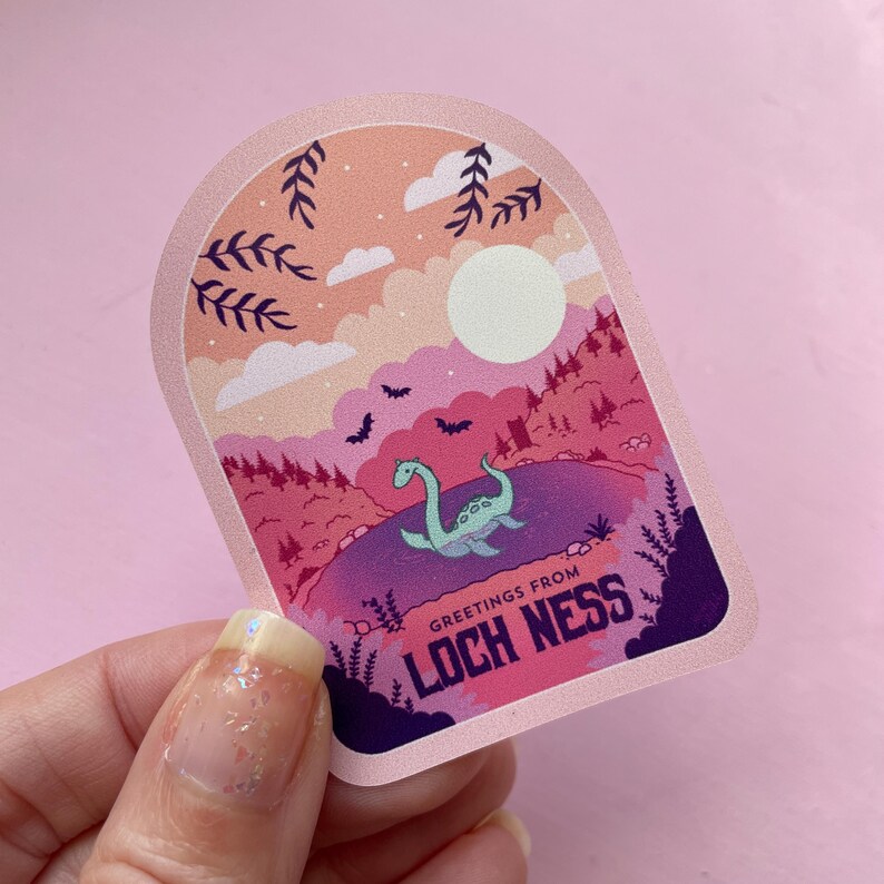 K&ouml;nnte beinhalten: Ein rosa und lilafarbener Aufkleber mit einer Cartoon-Illustration eines gr&uuml;nen Seeungeheuers im Loch Ness in einem See. Der Aufkleber hat einen rosa Bogen mit dem Text "Greetings from Loch Ness" in Wei&szlig;.