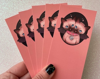 May include: A stack of pink bookmarks featuring a Siamese cat with two heads, bat wings, and a pink collar with a heart-shaped tag. The cat is set against a dark background with stars. The bookmarks are held in a hand with a black and gold nail design.