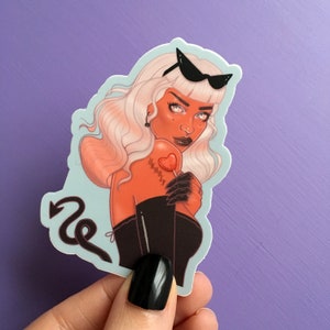 K&ouml;nnte beinhalten: Ein Aufkleber mit einer stilisierten Illustration einer d&auml;monischen Frau mit wei&szlig;en Haaren, roter Haut und schwarzen Accessoires. Sie h&auml;lt einen herzf&ouml;rmigen Lutscher und hat das Wort "Unholy" auf ihrer Brust t&auml;towiert. Ein Teufelsschwanz ist ebenfalls vorhanden.