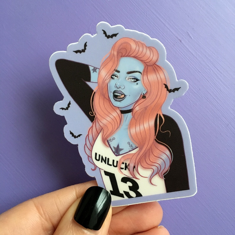 K&ouml;nnte beinhalten: Ein Aufkleber mit einer stilisierten Illustration einer Frau mit blauer Haut, rosa Haaren und einem schwarzen Halsband. Die Frau tr&auml;gt ein wei&szlig;es Tanktop mit dem Text "UNLUCK 13". Schwarze Flederm&auml;use sind um das Bild verstreut.