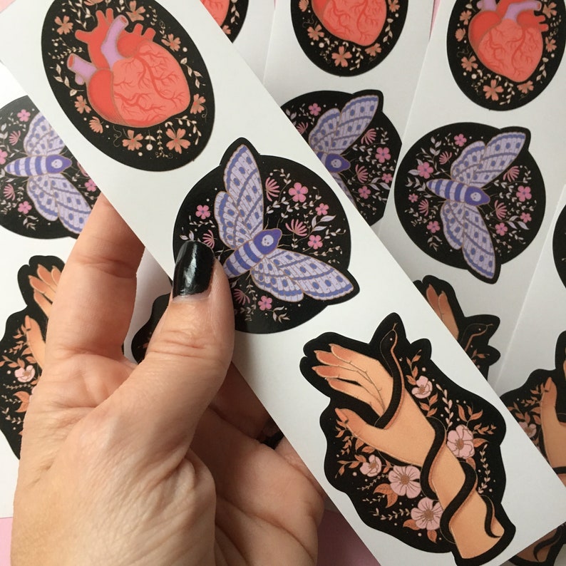 May include: A sheet of stickers featuring various designs. One sticker shows a human heart with floral accents. Another displays a moth with purple wings and a floral background. A third sticker depicts a hand holding a snake, surrounded by flowers.