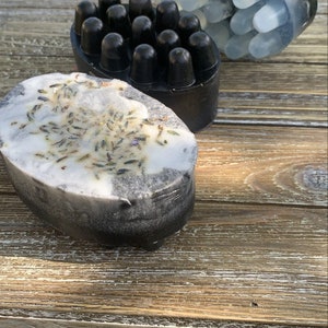 May include: Three black and white soap bars. One bar has a textured surface with small bumps. Another bar has a smooth surface with a white layer and dried lavender flowers. The third bar has a smooth surface with a clear layer.