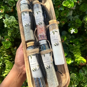 May include: A wooden tray holds six glass bottles of bath salts, each sealed with a metal cap and tied with twine. The bottles contain various colors of salts, and each has a label that reads "Relaxing Bath Salt". The background is green foliage.
