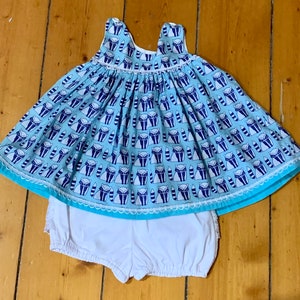 以下が含まれることがあります： 白いトリムとアライグマのパターンが付いた、薄いブルーのノースリーブドレス。ドレスは白いブルマと組み合わされています。