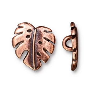 May include: A pair of copper-colored metal buttons shaped like a monstera leaf. The buttons have a loop on the back for sewing.