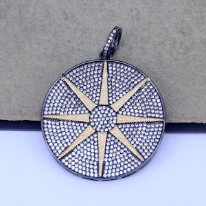 May include: A round pendant with a black metal frame and a starburst design. The starburst is made of small white stones with gold accents.