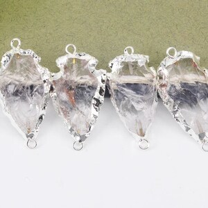 May include: Four clear quartz arrowhead pendants with silver-colored metal edging and loops for hanging. The quartz has natural inclusions and variations in color. The pendants are arranged on a white surface with a green and black background.