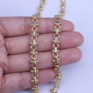 Op de afbeelding: Een gouden ketting met een delicaat, ingewikkeld ontwerp. De ketting is gemaakt van kleine, ronde schakels die met elkaar verbonden zijn door een reeks kleinere, vierkante schakels. De ketting is ongeveer 45 cm lang.