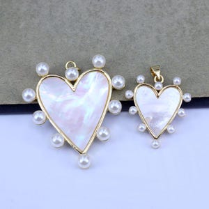 Peut inclure: Deux pendentifs en forme de cœur dorés avec des accents de perles blanches. Le plus grand pendentif a un incrustation de nacre blanche. Les deux pendentifs ont une boucle en haut pour s'attacher à une chaîne.