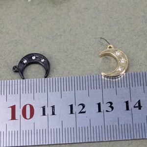 Pode incluir: Dois pingentes em forma de lua crescente com um design de estrela. O pingente &agrave; esquerda &eacute; preto com strass transparentes. O pingente &agrave; direita &eacute; dourado com strass transparentes. Ambos os pingentes s&atilde;o medidos com uma r&eacute;gua com marcas de 10 a 14.