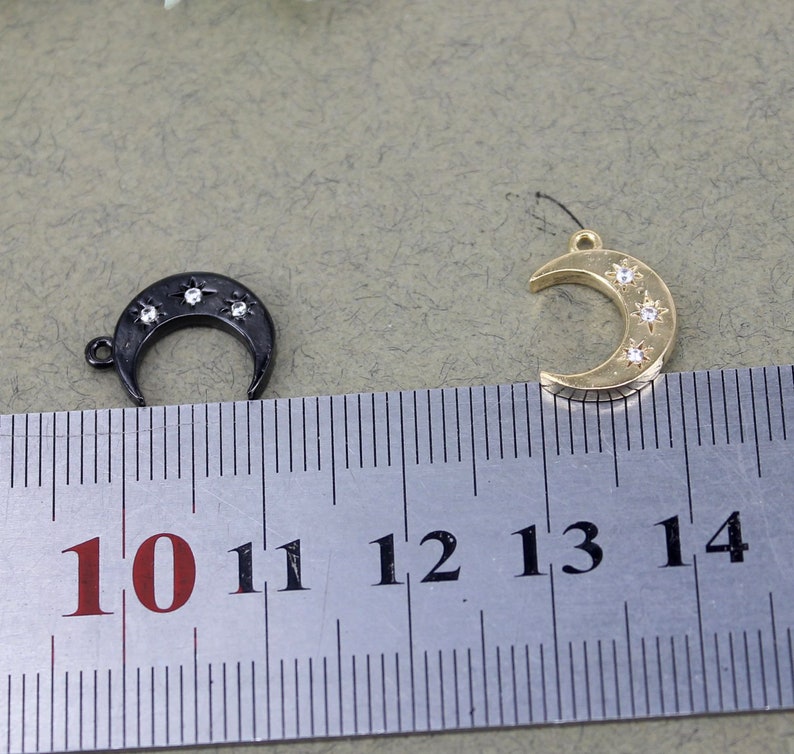 Pode incluir: Dois pingentes em forma de lua crescente com um design de estrela. O pingente &agrave; esquerda &eacute; preto com strass transparentes. O pingente &agrave; direita &eacute; dourado com strass transparentes. Ambos os pingentes s&atilde;o medidos com uma r&eacute;gua com marcas de 10 a 14.
