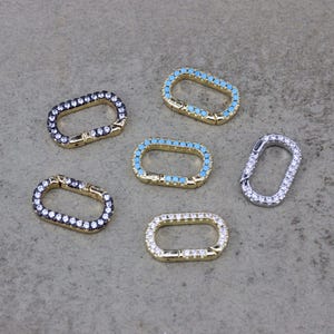 May include: A collection of oval-shaped jewellery clasps. Some clasps are gold-toned with clear or black gemstones, while others feature turquoise-coloured stones. One clasp is silver-toned with clear gemstones. The clasps are on a grey surface.