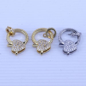 Könnte beinhalten: Drei herzförmige Verschlüsse mit einem runden Ring, jeder mit einer anderen Oberfläche: Gold, Silber und Rhodium. Die Verschlüsse sind mit klaren Zirkonia-Steinen verziert.