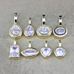 May include: A collection of eight gold-toned metal pendants, each featuring a clear, faceted gemstone. The pendants come in various shapes, including pear, oval, rectangular, marquise, heart, and round, all equipped with a top bail for hanging.
