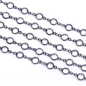 May include: A close-up of a black chain with clear round beads. The chain is laid out in a straight line on a white surface.