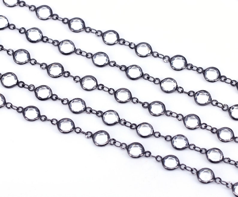 May include: A close-up of a black chain with clear round beads. The chain is laid out in a straight line on a white surface.
