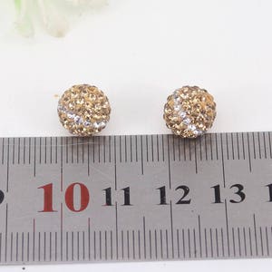 May include: Two gold-toned round earrings with clear and gold rhinestones. The earrings are about 1 cm in diameter and are shown next to a ruler.