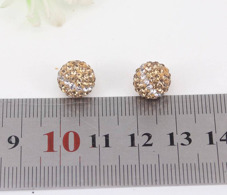 May include: Two gold-toned round earrings with clear and gold rhinestones. The earrings are about 1 cm in diameter and are shown next to a ruler.