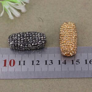 May include: Two oval-shaped rhinestone beads, one black and one gold, measuring approximately 1 inch in length. The beads are shown next to a ruler with markings from 10 to 16.