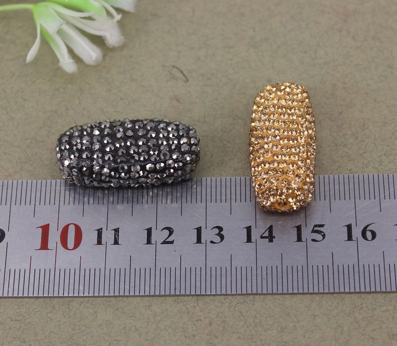 May include: Two oval-shaped rhinestone beads, one black and one gold, measuring approximately 1 inch in length. The beads are shown next to a ruler with markings from 10 to 16.