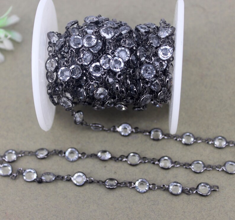 May include: A roll of black chain with clear faceted glass beads. The chain is on a white spool and there are three strands of the chain laid out on a gray surface.