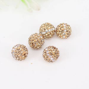 May include: Six gold-toned round beads with clear rhinestones. The beads are scattered on a white surface.