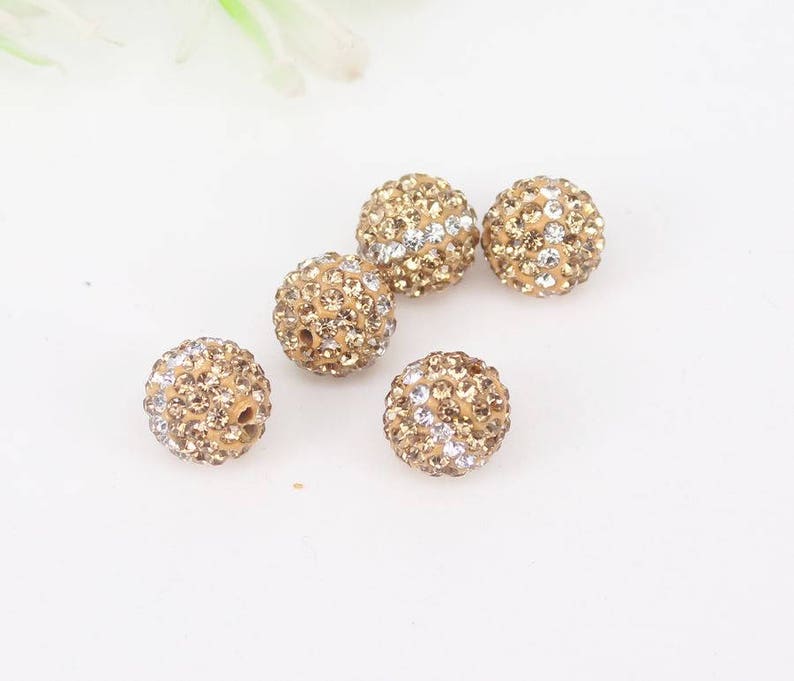 May include: Six gold-toned round beads with clear rhinestones. The beads are scattered on a white surface.