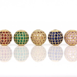 Pode incluir: Cinco contas redondas com engastes de metal dourado e detalhes de cristal coloridos. As contas são vermelhas, verdes, castanho claro, azuis e cor-de-rosa.