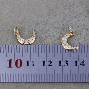 May include: Two gold crescent moon charms with white opal stones and clear rhinestones. The charms are approximately 1.25 inches in length and are measured against a ruler.