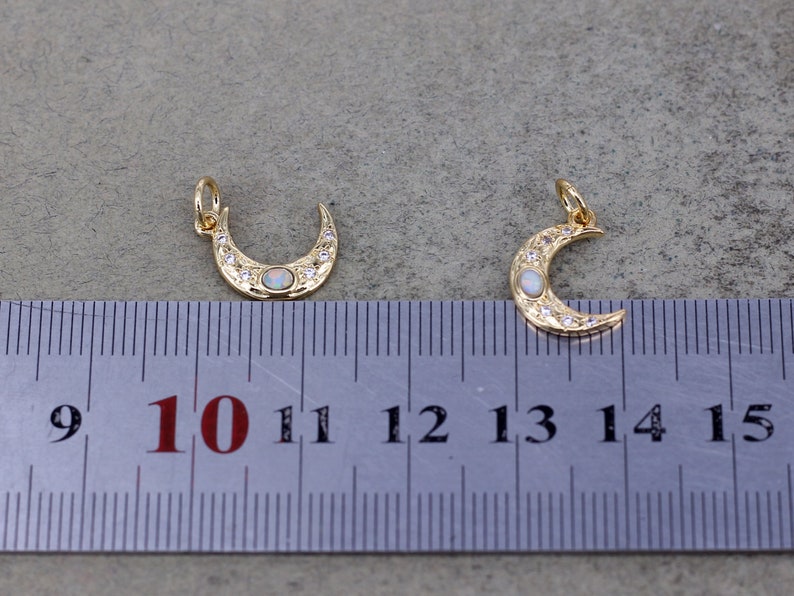 May include: Two gold crescent moon charms with white opal stones and clear rhinestones. The charms are approximately 1.25 inches in length and are measured against a ruler.