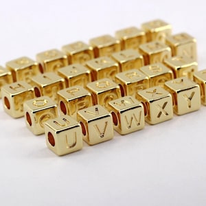 May include: Gold-colored alphabet beads, each cube-shaped with a letter from the alphabet engraved on one side. The beads have a shiny finish and are arranged in rows on a white surface. Ideal for jewelry making.