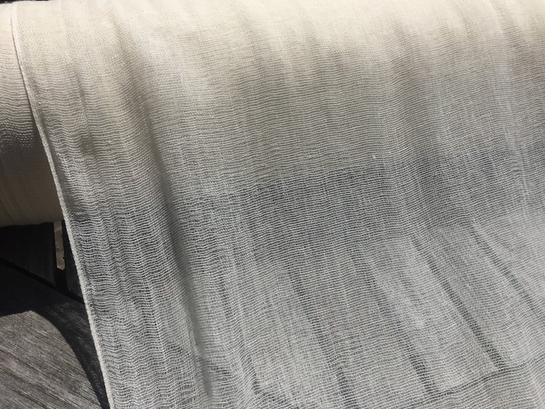 May include: A roll of white sheer fabric with a fine mesh texture. The fabric is draped over a wooden surface.