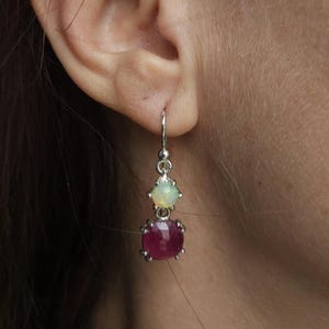May include: A pair of silver drop earrings, each featuring a vibrant, cushion-cut ruby gemstone below a smaller, oval-shaped opal. The earrings are suspended from a simple hook design, showcasing the colorful gemstones.