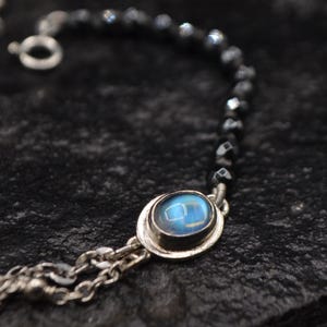 Puede incluir: Un collar de plata con un colgante de piedra azul y una cadena de cuentas negras. El colgante ovalado está engastado en un bisel de plata. El collar está sobre una superficie oscura y texturizada.