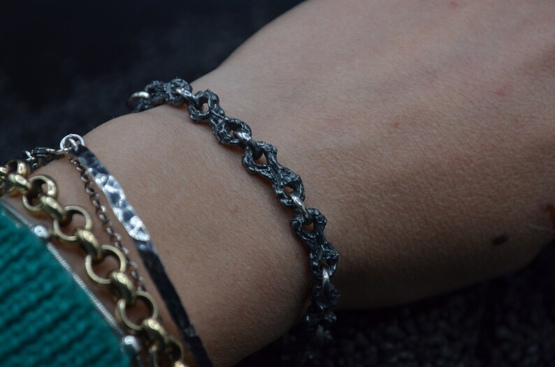 May include: A silver bracelet with a textured, chain-link design. The bracelet is worn on a person's wrist.